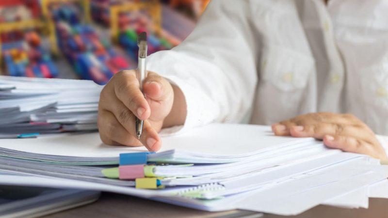 Businessman hands holding pen for working in Stacks of paper files searching information business report papers and piles of unfinished documents achieves on laptop computer desk in modern office